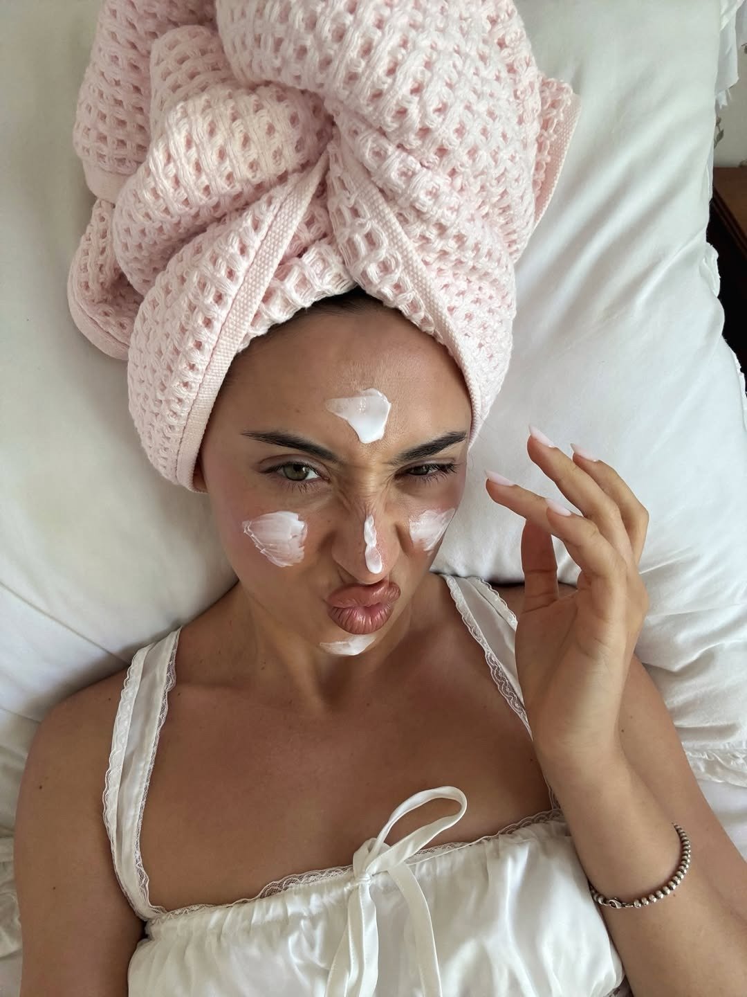 Ácido mandélico para que serve: modelo posa na cama com toalha rosa texturizada, camisola branca e creme no rosto, clima leve