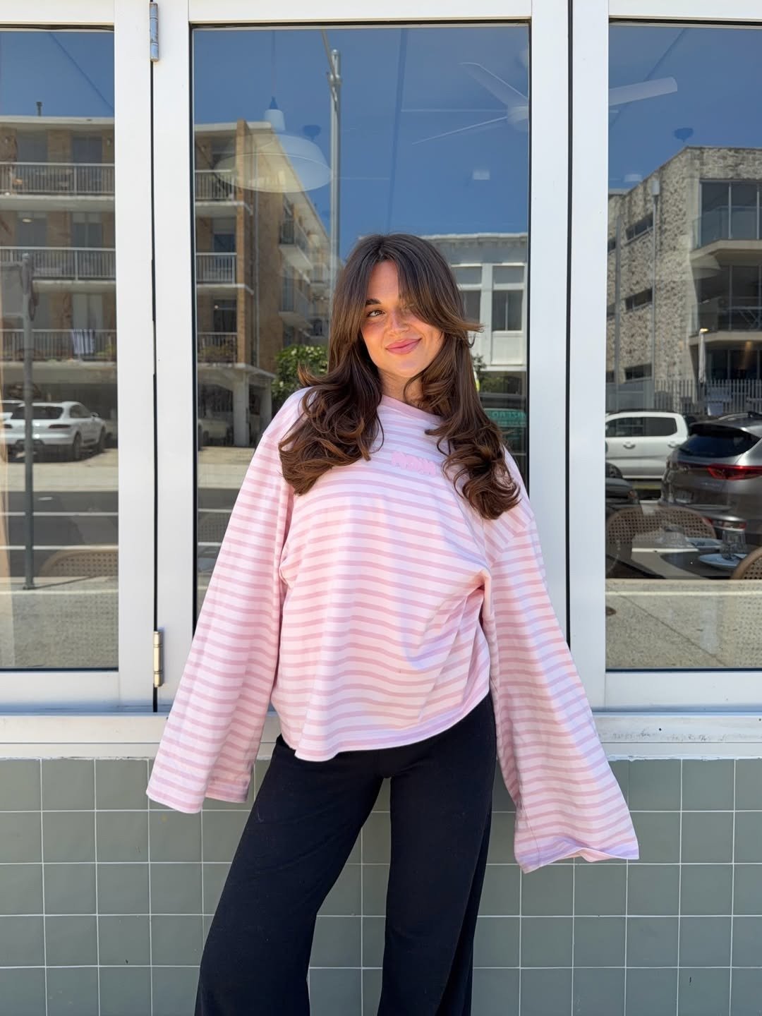 O que é sulfato no shampoo: modelo posa com blusa listrada rosa de mangas amplas e calça preta diante de janela urbana leve.