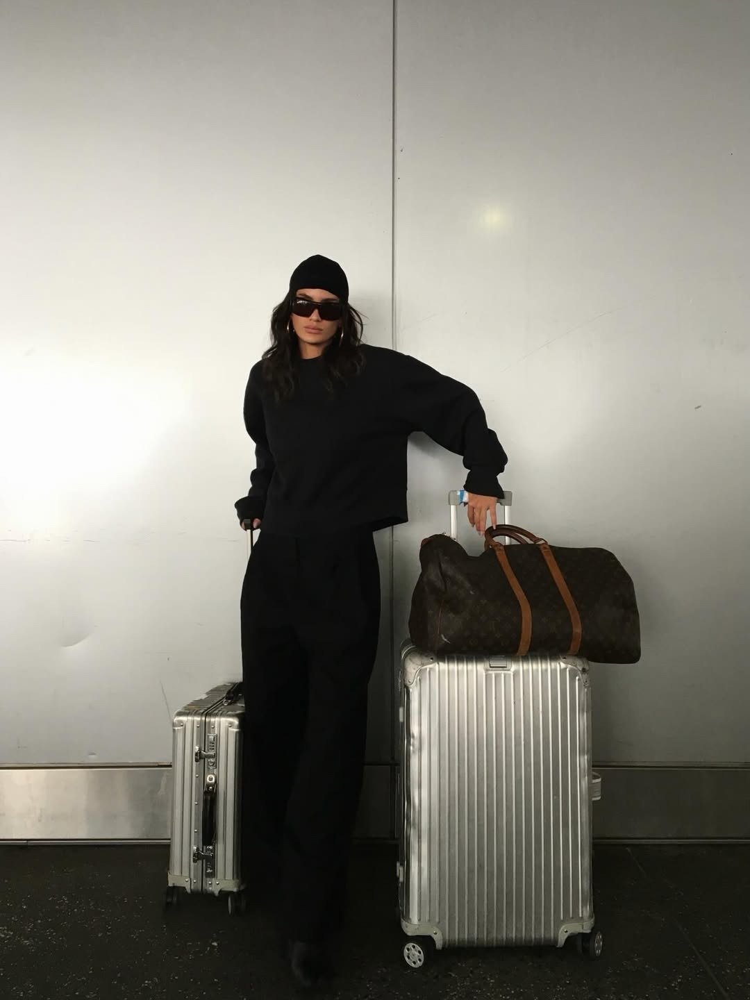 Modelos de calças de alfaiataria: modelo posa de preto com caimento amplo, mala prateada e clima sóbrio urbano.