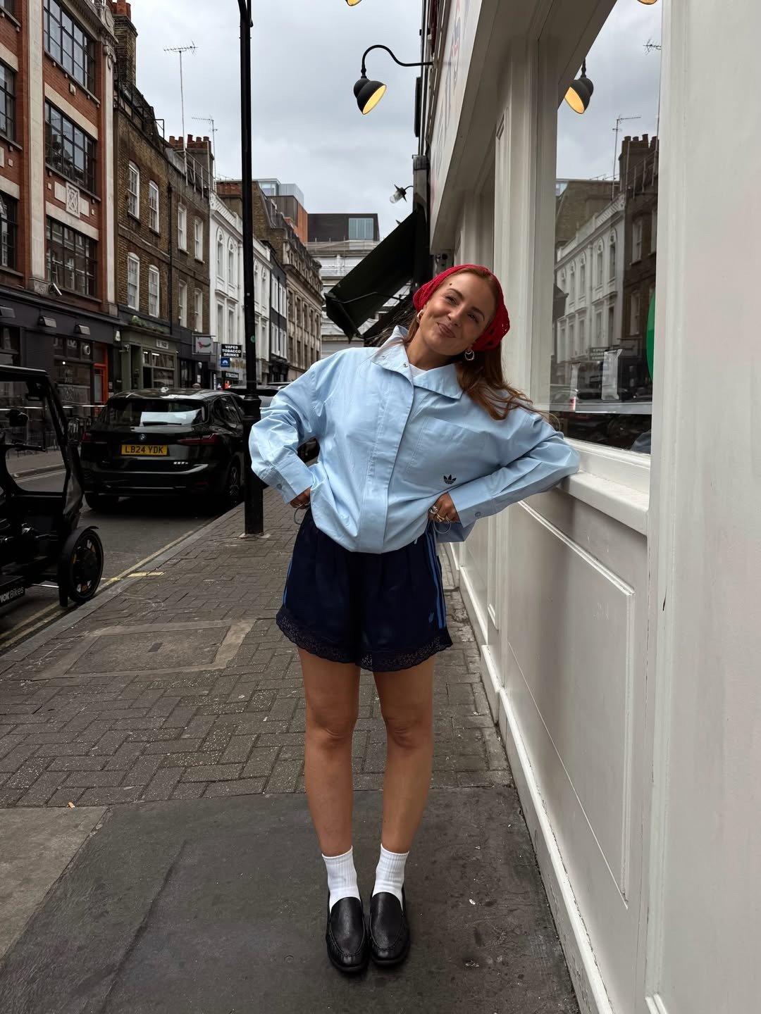 Modelo posa sorrindo na rua com jaqueta azul ampla, short escuro e loafers pretos com meia branca, entre os melhores sapatos