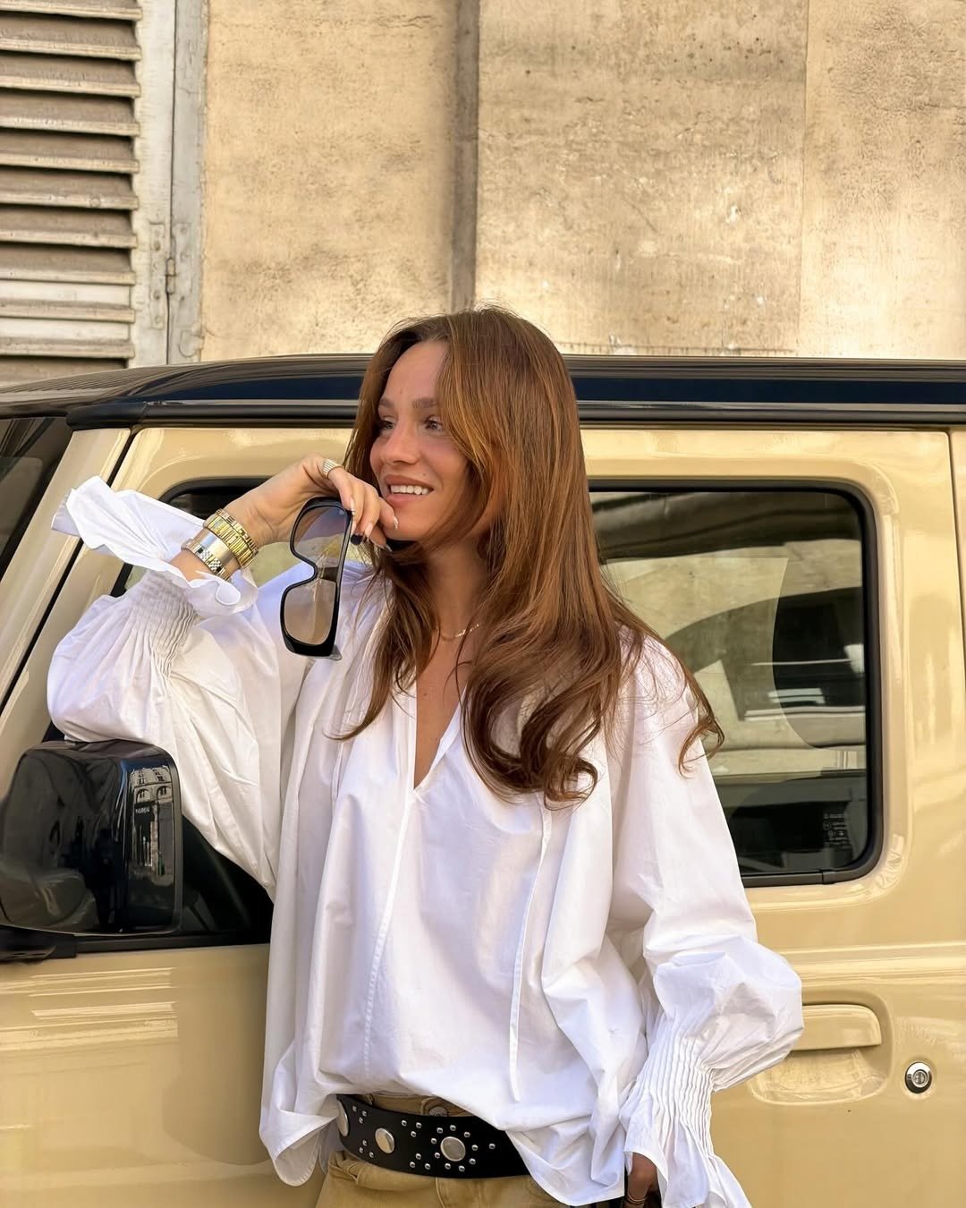 O que é sulfato no shampoo: modelo posa sorrindo com blusa branca ampla, cinto preto e carro bege em clima elegante.