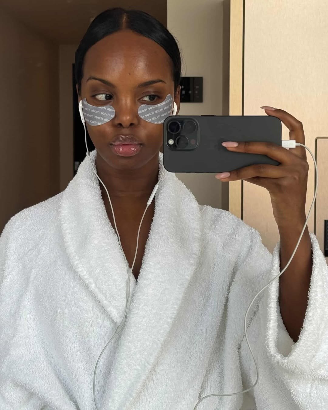 Modelo posa de roupão branco felpudo com patches nos olhos e celular no espelho, clima clean de skincare e melhor sabonete