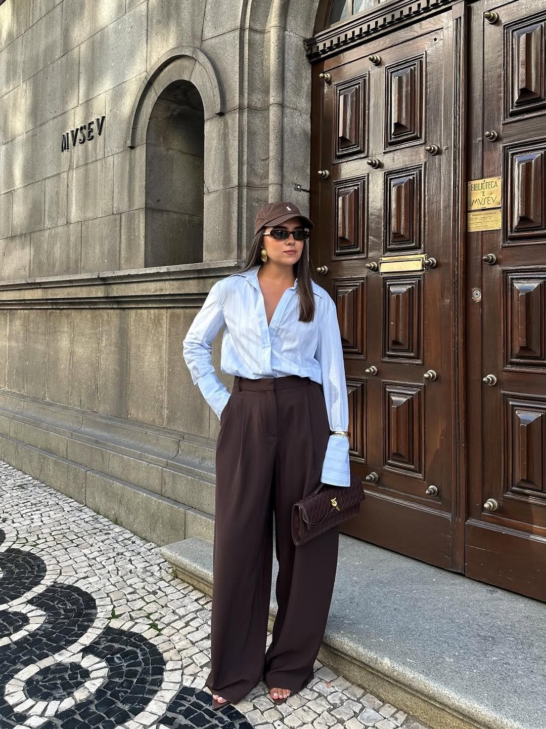 Modelos de calças de alfaiataria: modelo posa com pantalona marrom e camisa azul clara diante de porta clássica.