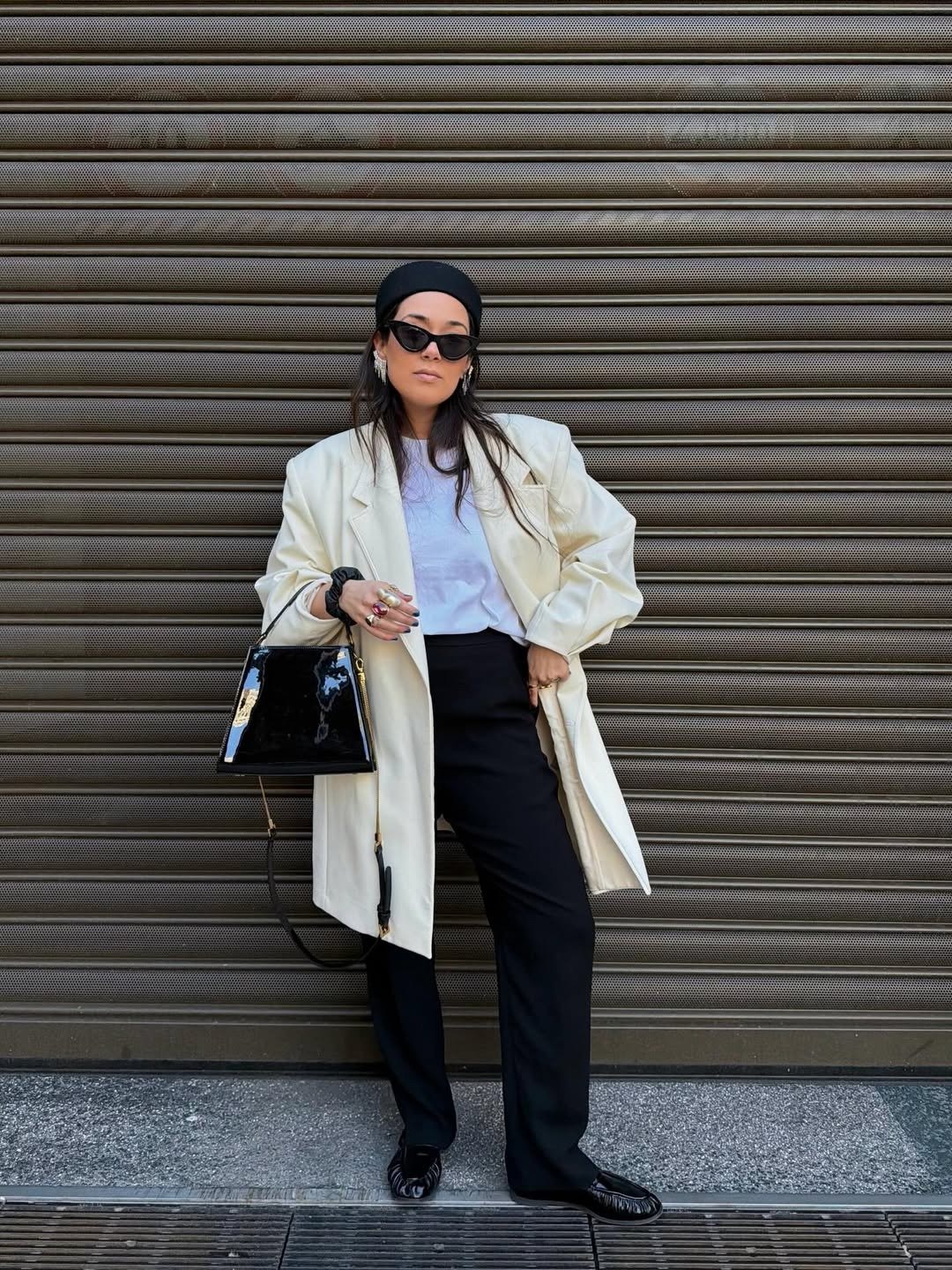 Modelos de calças de alfaiataria: modelo posa com calça preta reta, blazer off-white e bolsa verniz em clima urbano elegante.