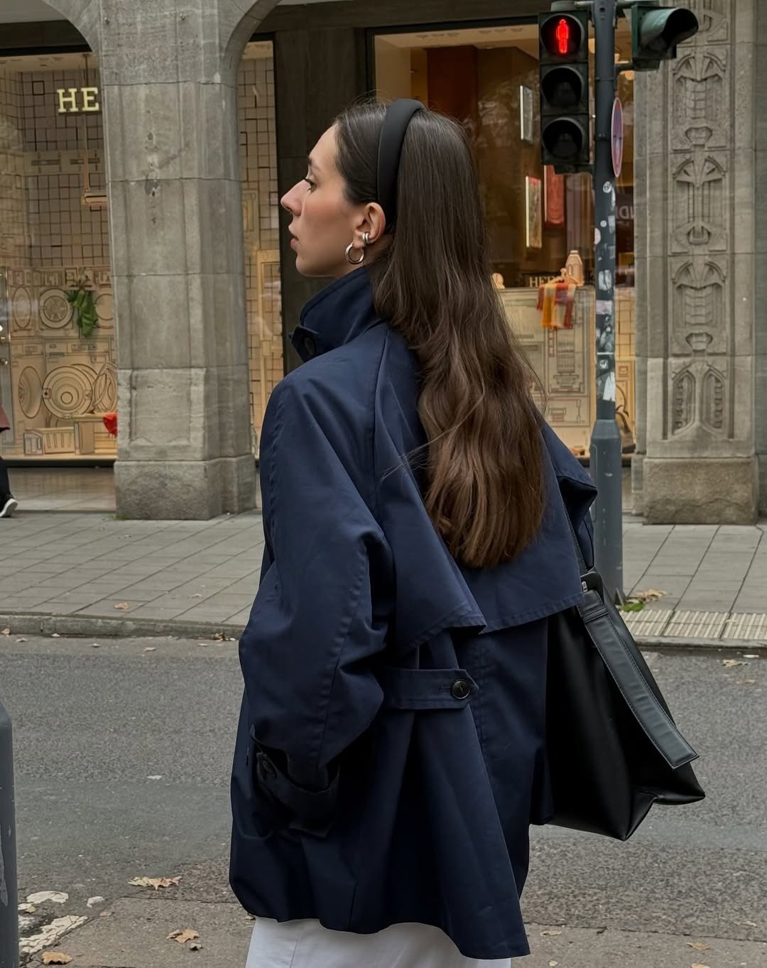 Acessórios para o outono: modelo posa de perfil com tiara, argolas e bolsa preta, trench azul-marinho em clima urbano sóbrio