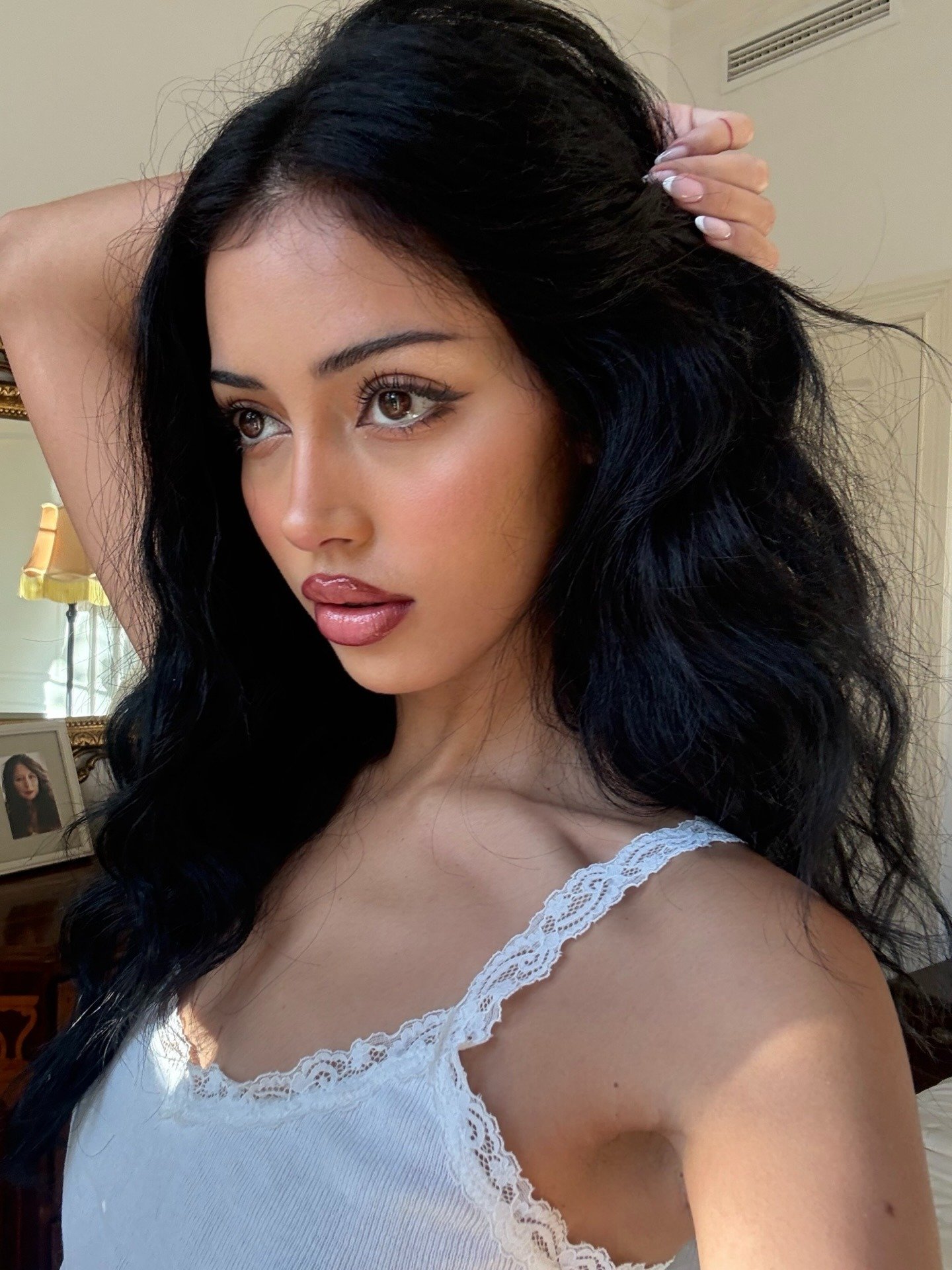 Modelo posa com sobrancelha fina, regata branca canelada com renda e cabelo preto ondulado em clima elegante e suave