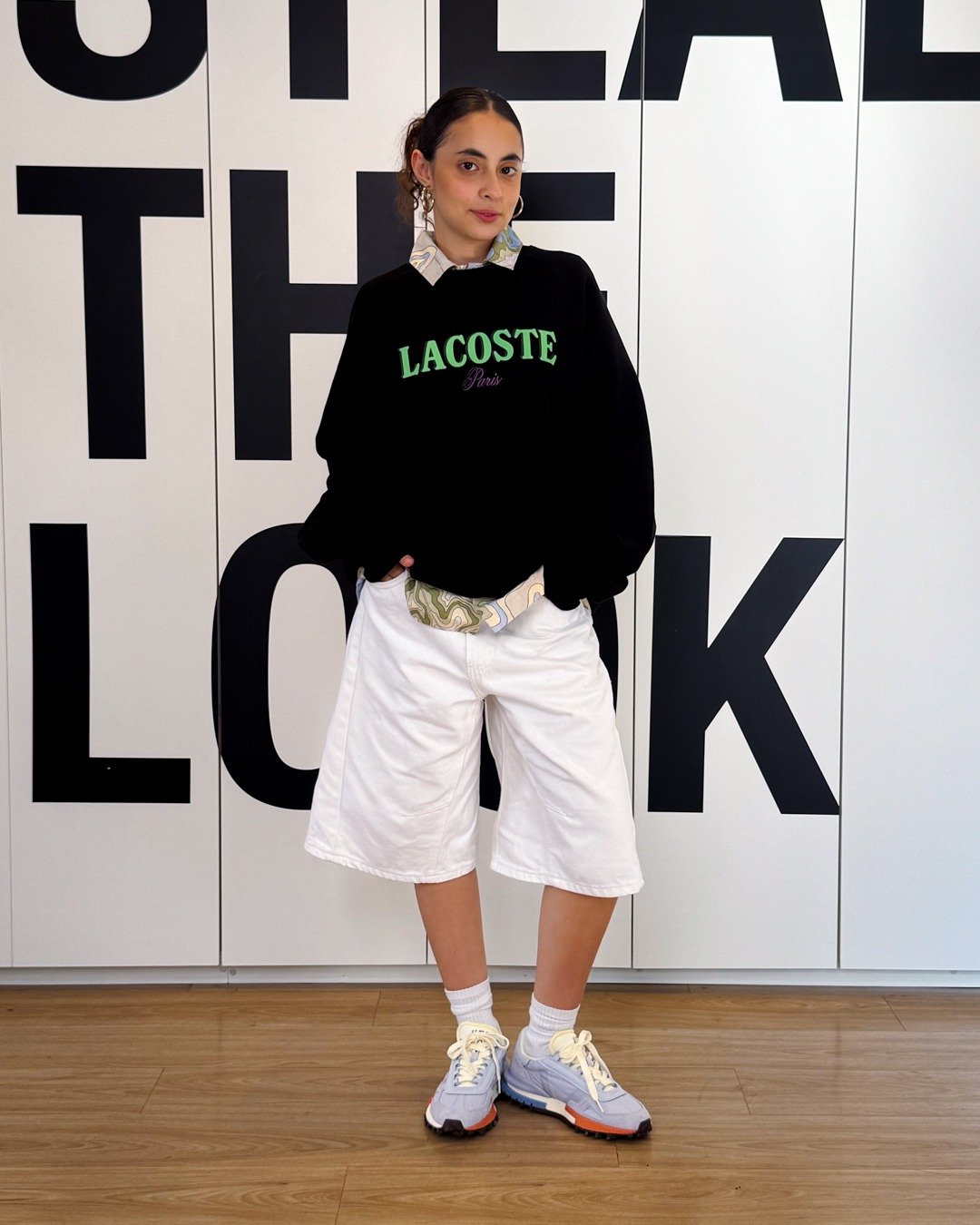 Modelo posa com tênis versátil e estiloso, moletom preto, bermuda branca ampla e camisa estampada; clima casual.