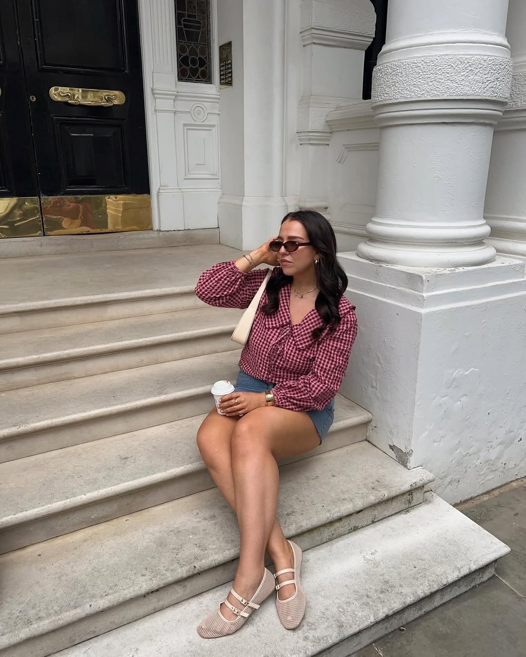 Modelo posa sentada na escada com sapatilha de tela bege, blusa xadrez vinho e short jeans em clima elegante.