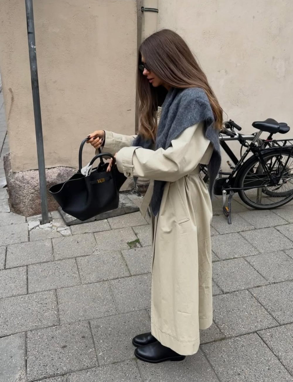 Tendência clássica: modelo posa com trench coat bege, tricô cinza e bolsa preta em rua urbana, clima elegante.