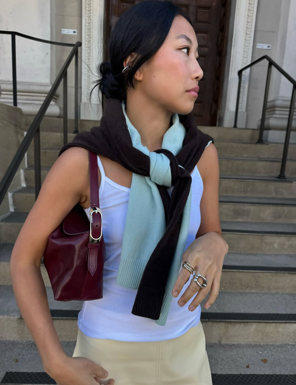 Tendência clássica: modelo posa de perfil com regata branca, malha nos ombros, saia clara e bolsa vinho em clima elegante.