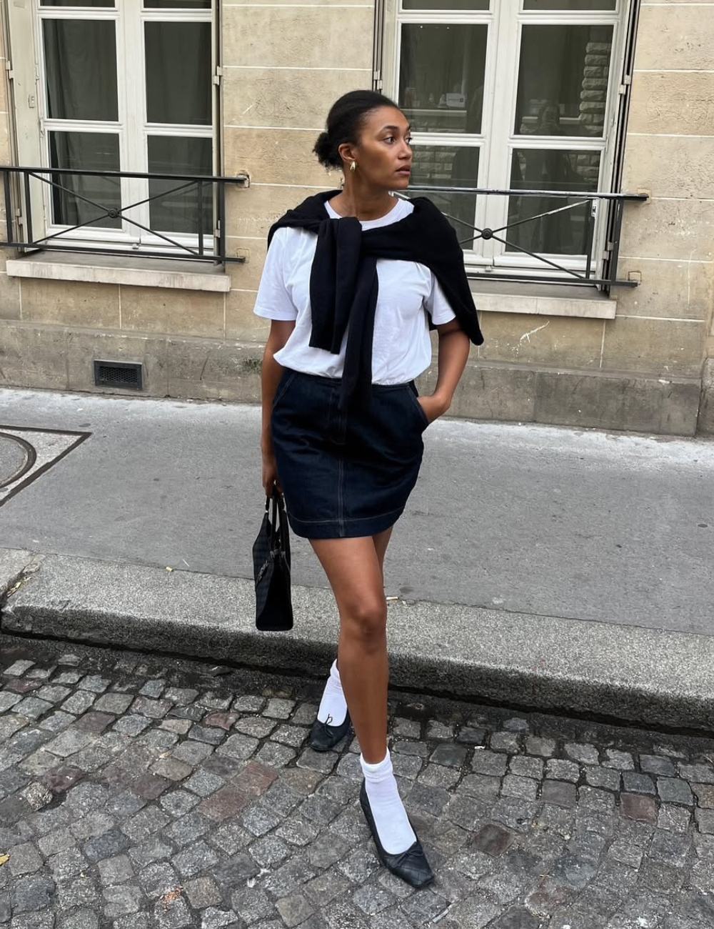 Tendência clássica: modelo posa com camiseta branca, suéter preto nos ombros e minissaia escura em rua serena.