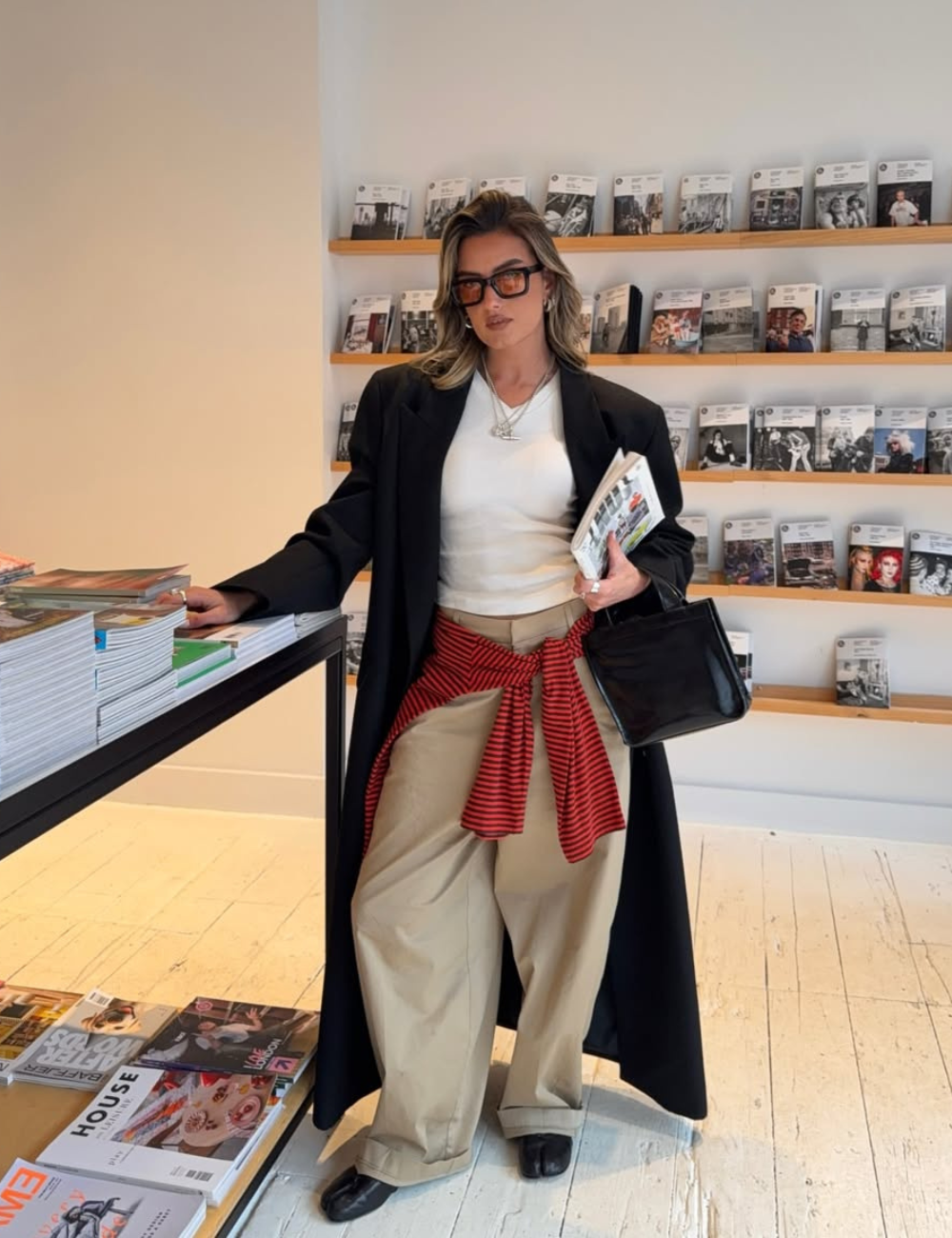 Tendência clássica: modelo posa com blazer preto, camiseta branca, calça bege ampla e tricô listrado em livraria elegante.