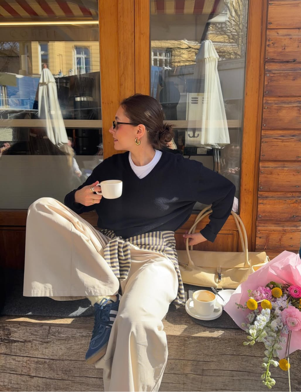 Tendência clássica: modelo posa com tricô preto, calça ampla creme e tênis azul em café ensolarado, clima elegante e sereno