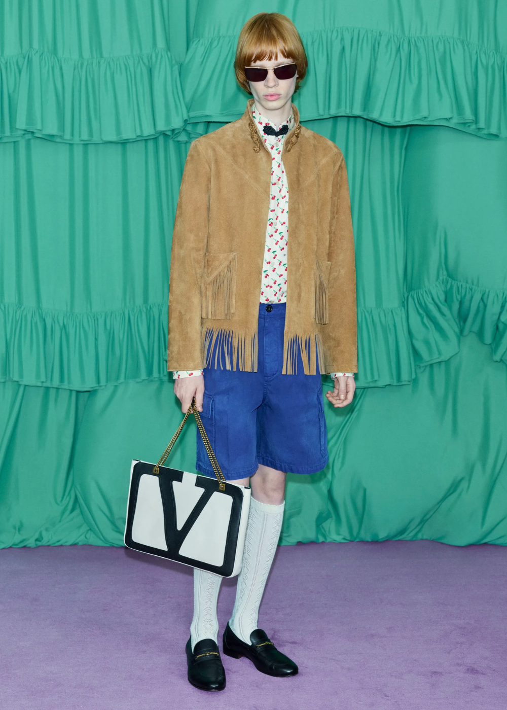 Looks com franja: modelo posa com jaqueta camel de camurça, bermuda azul e bolsa V em cenário vibrante.