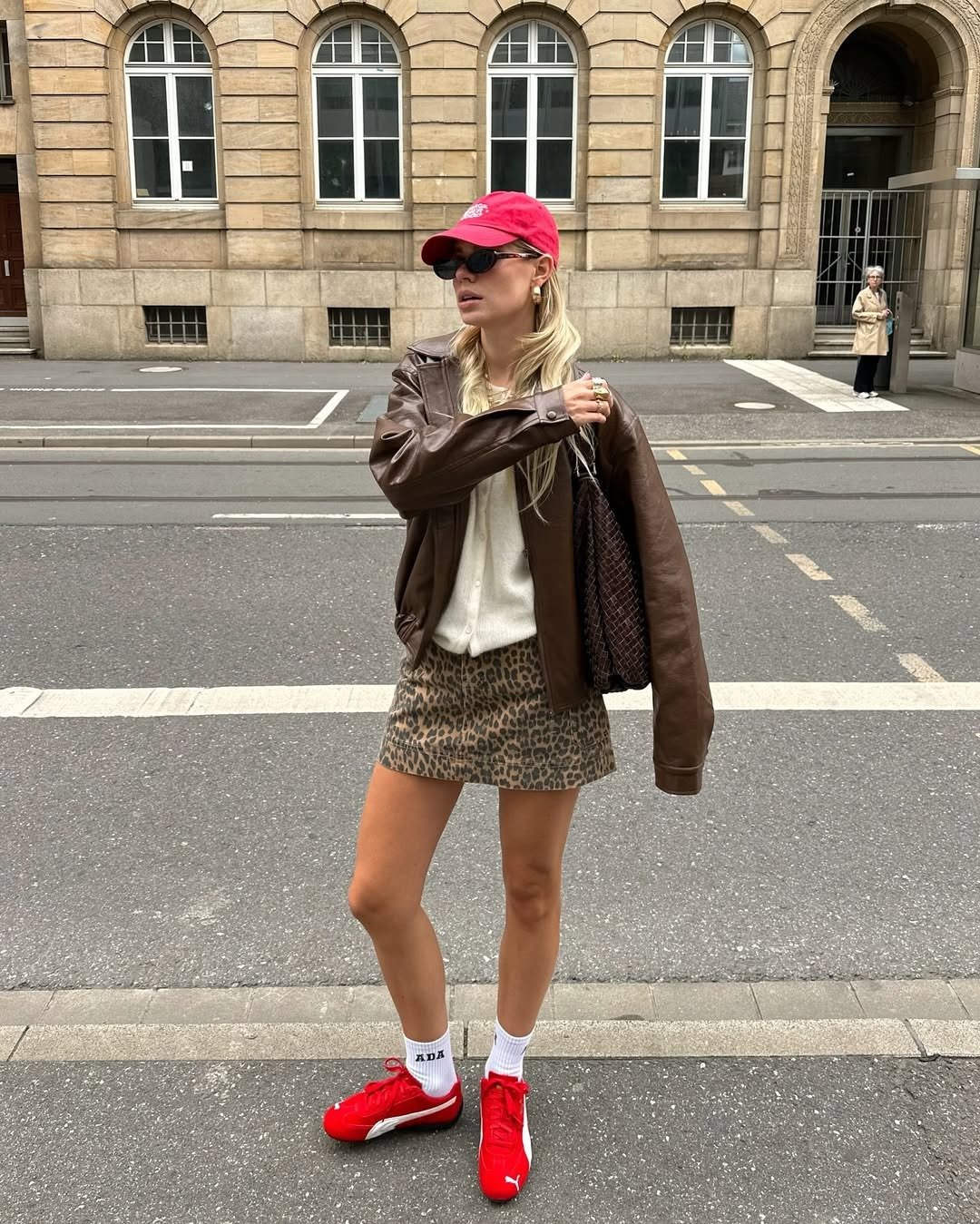 Modelo posa na rua com tênis da Puma vermelhos, jaqueta de couro marrom, minissaia animal print e ar urbano casual.
