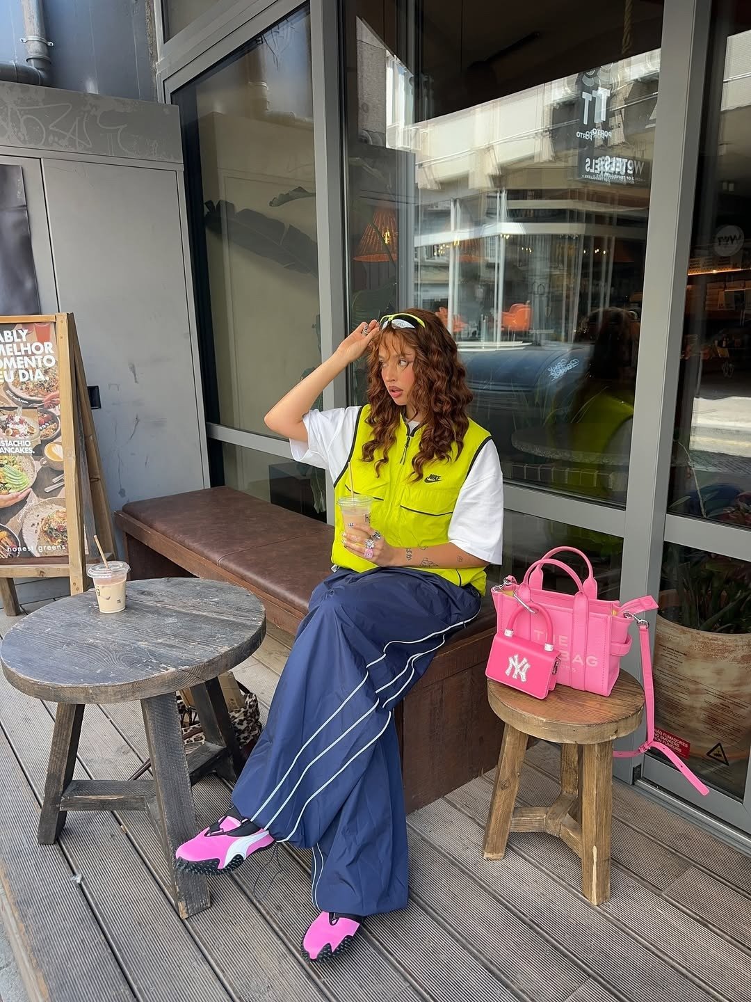 Modelo posa sentada com tênis da Puma rosa, colete neon, calça ampla azul e bolsa pink em café urbano despojado.