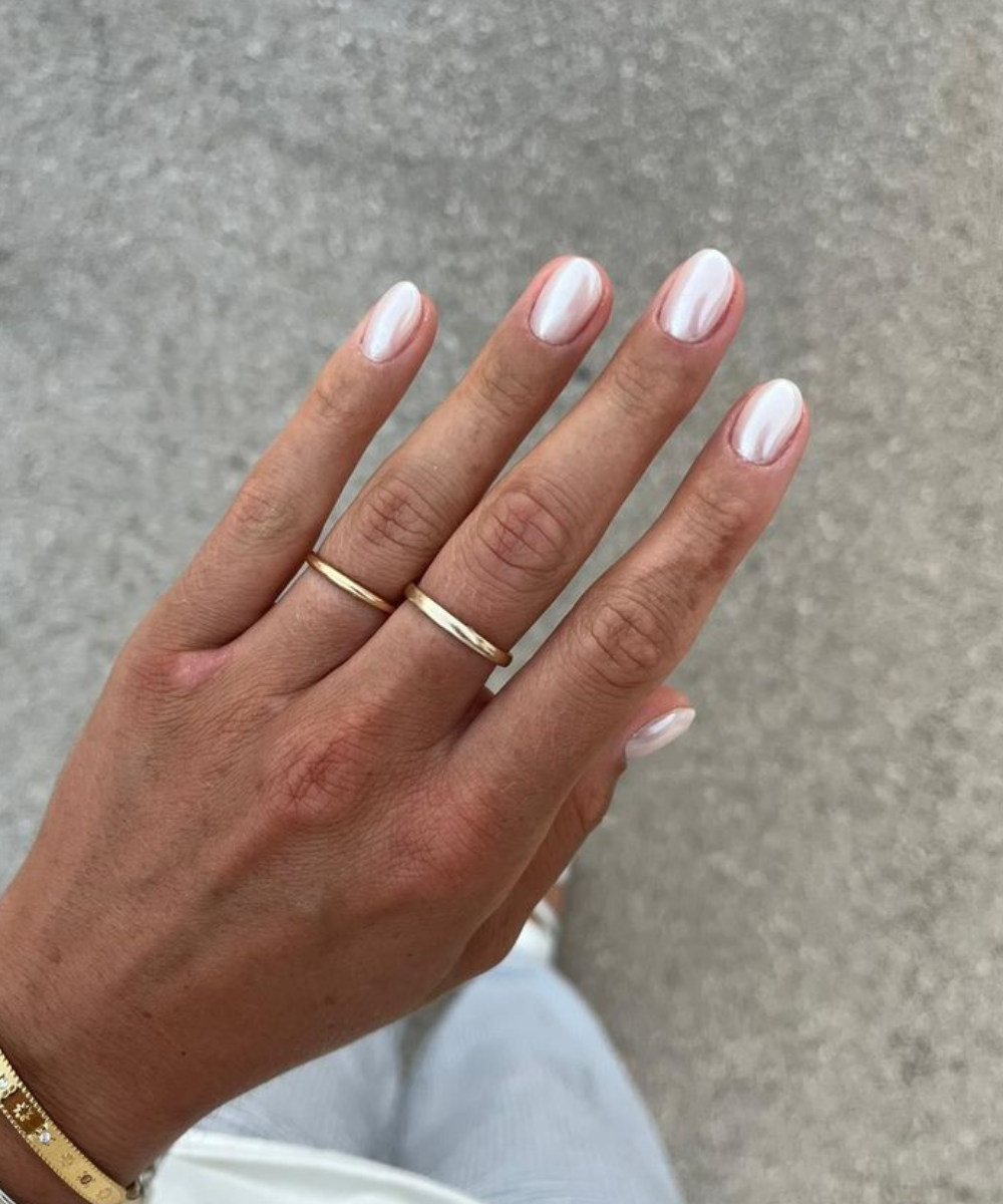 Unhas postiças peroladas em formato oval brilham na mão com anéis dourados, em close minimalista e elegante.