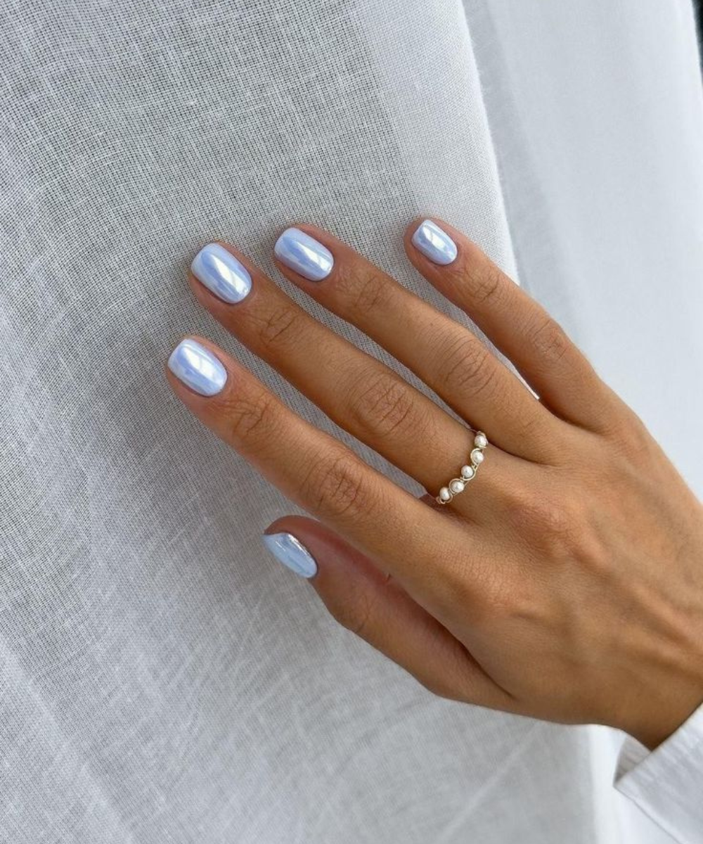 Unhas postiças curtas azul-perolado brilham em mão relaxada com anel de pérolas sobre tecido branco, clima delicado.