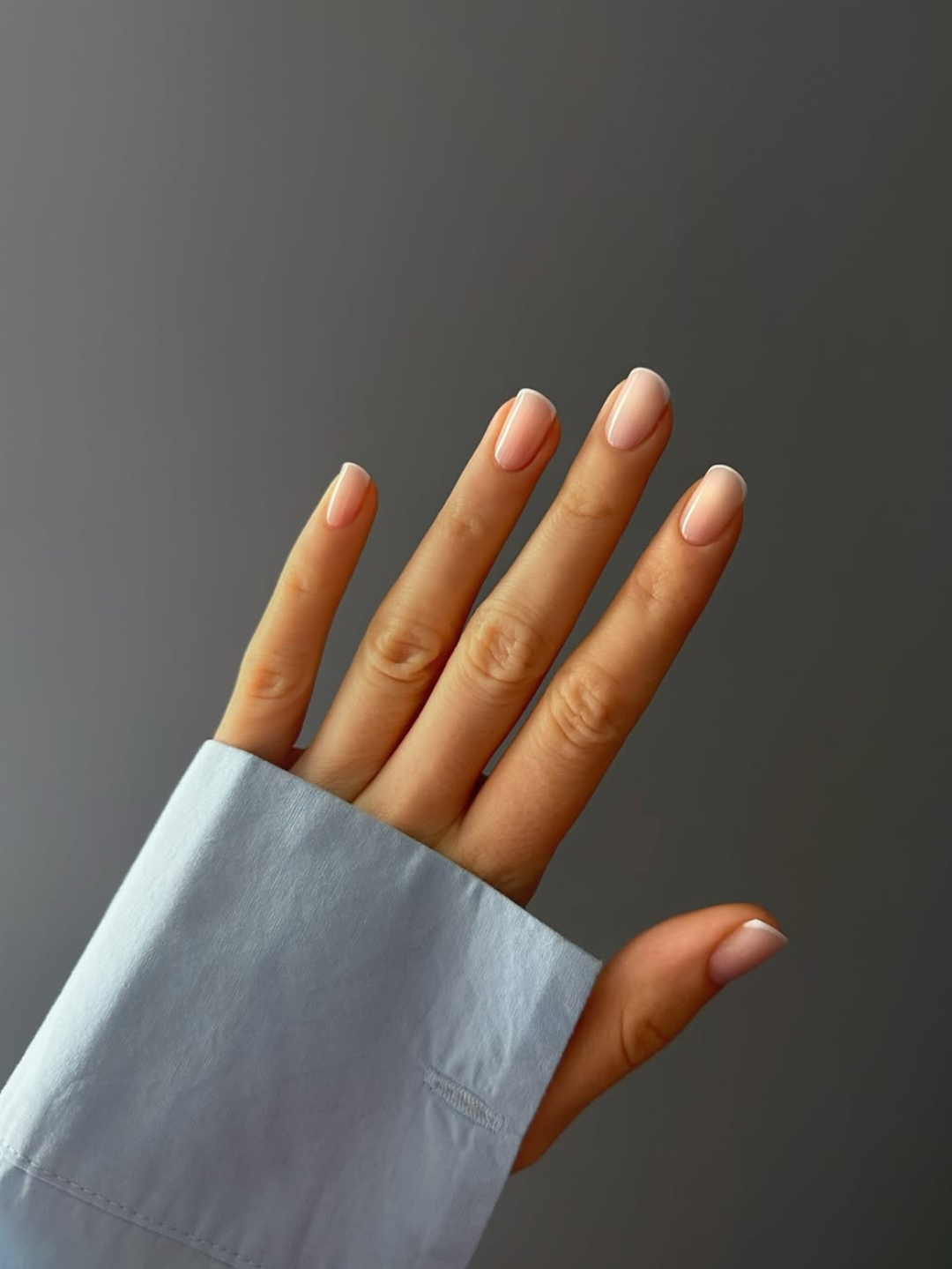 Unhas para madrinha de casamento em nude rosado e formato curto oval, com manga azul-clara e fundo cinza elegante.