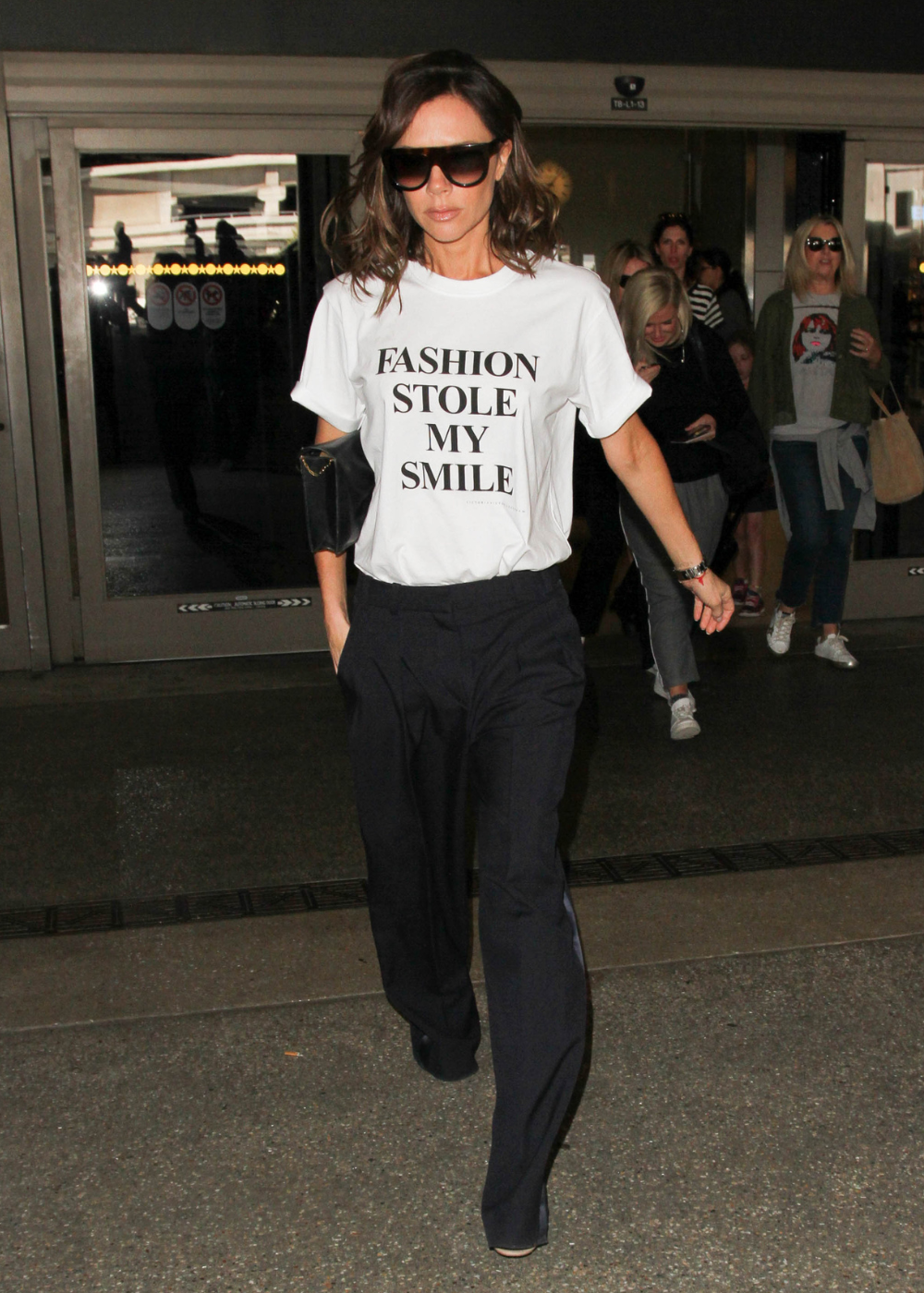 Modelo posa caminhando com camiseta branca oversized, calça preta e óculos escuros em clima urbano elegante.