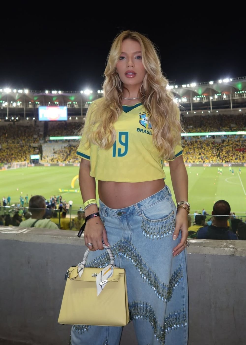 Modelo posa no estádio com cropped amarelo do Brasil, jeans claro bordado e bolsa estruturada, em clima glamouroso.