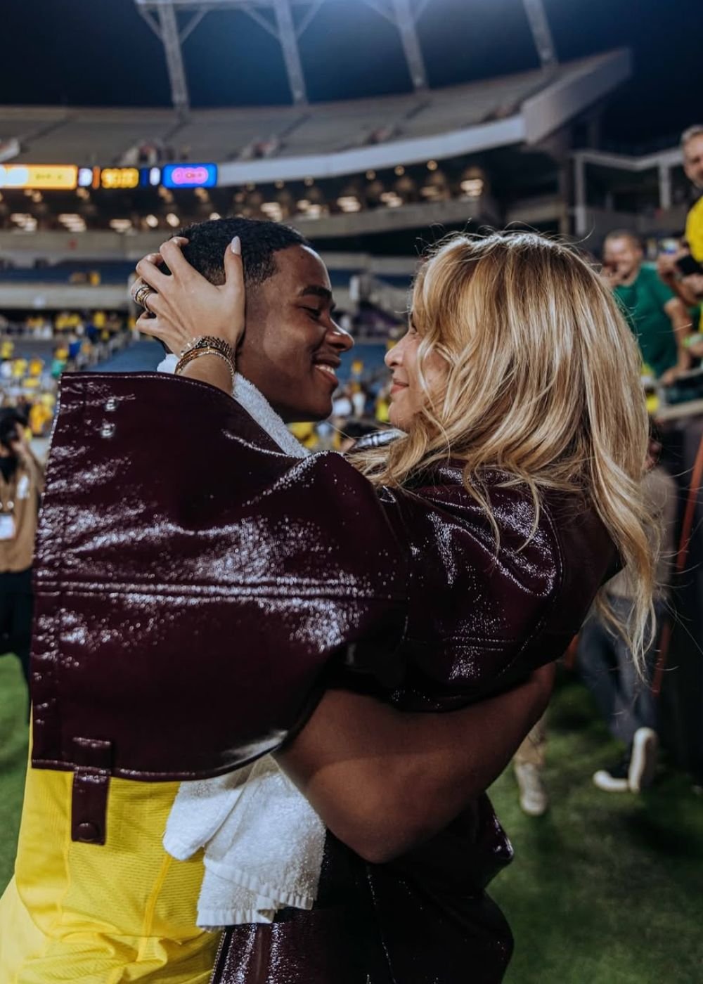 Casal se abraça no estádio; jaqueta vinho envernizada e uniforme amarelo criam clima romântico ligado ao universo WAG.