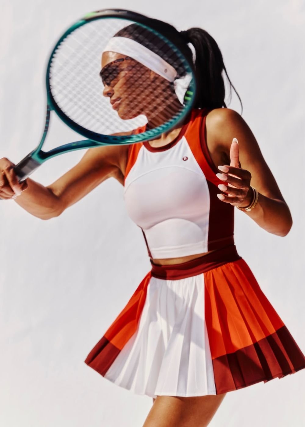 Tenista posa com top e saia plissada branca e laranja, viseira e raquete em cena clean, com energia esportiva.