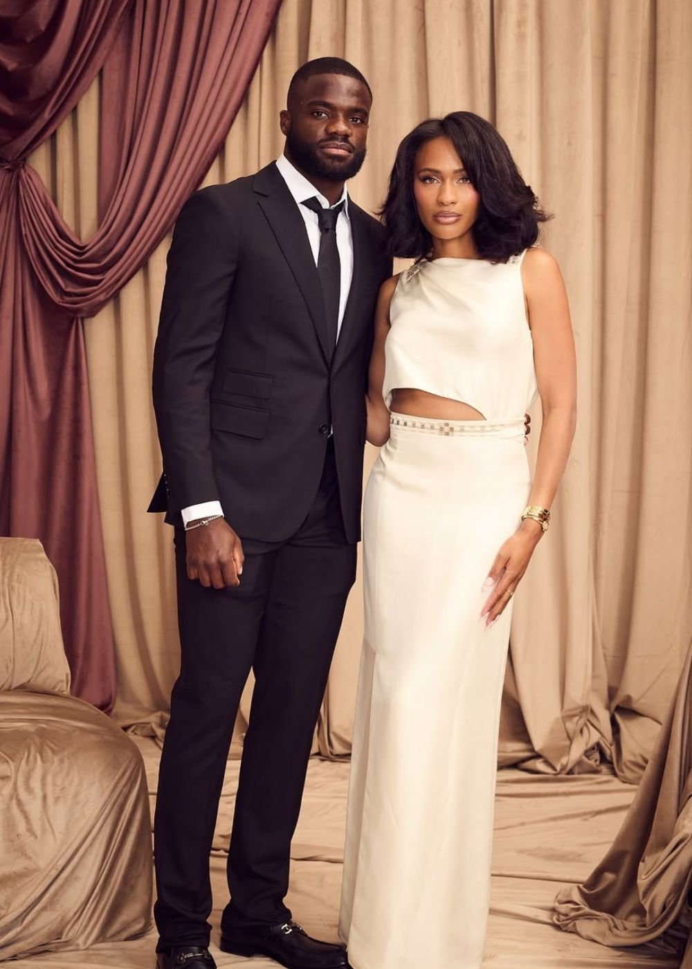 Casal posa elegante com terno preto e vestido off-white recortado em cetim, em cenário de cortinas bege sofisticadas