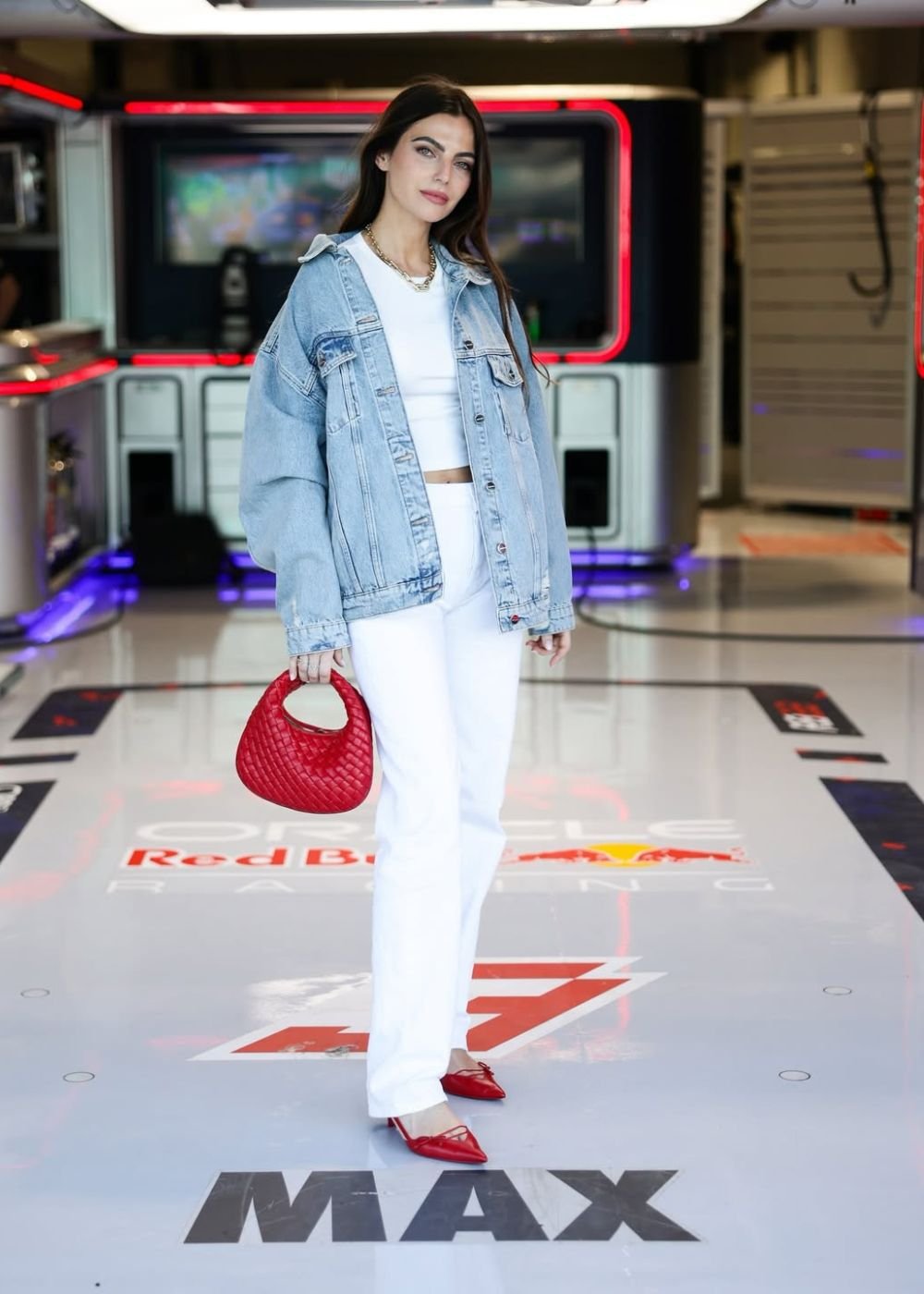 Modelo posa com jaqueta jeans oversized, look branco e bolsa vermelha em paddock glamouroso. Texto: Red Bull Racing MAX