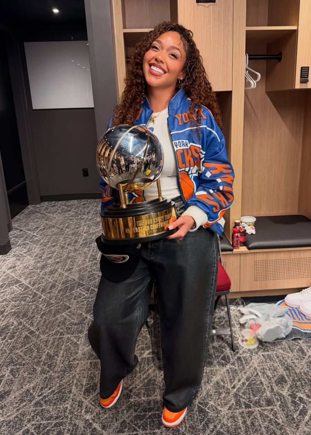 Afinal, o que é WAG? Mulher posa sorrindo com jaqueta esportiva azul e laranja dos Knicks e troféu. Texto: YORK CKS