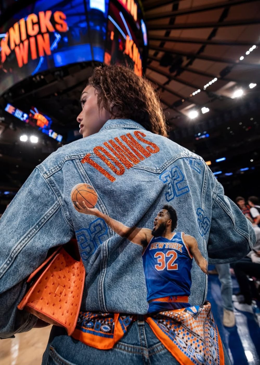 Modelo posa de costas na quadra com jaqueta jeans customizada azul e laranja, luxo esportivo. Texto: KNICKS WIN TOWNS 32 NEW