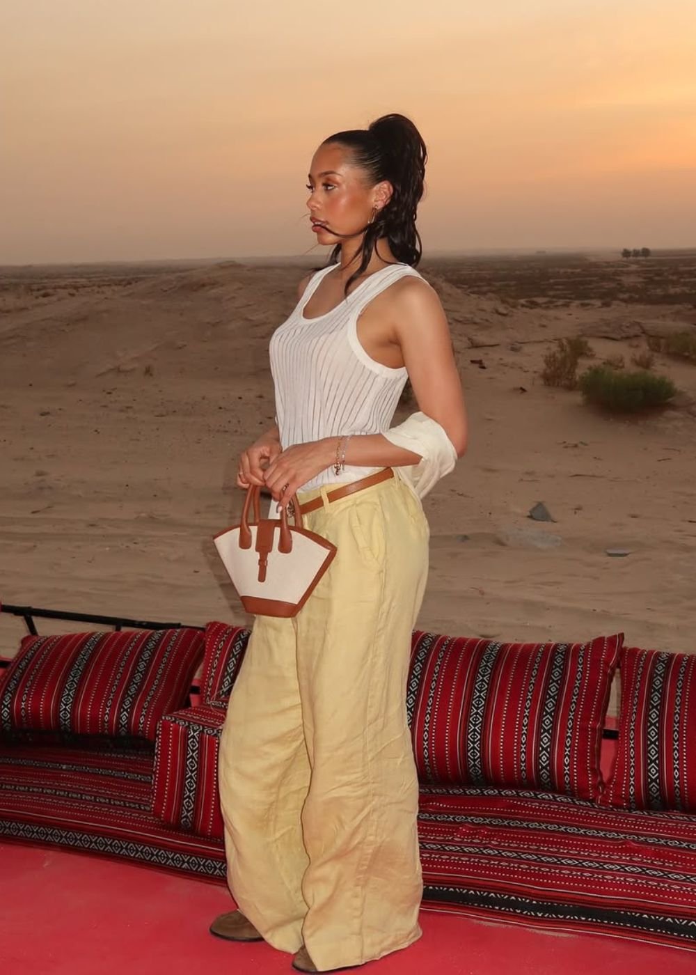 Modelo posa no deserto ao pôr do sol com regata canelada branca, calça ampla bege e bolsa bicolor, clima sofisticado.