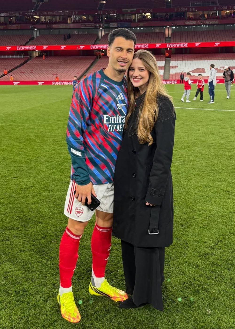 Casal posa sorrindo no gramado; jogador usa uniforme Arsenal vermelho e azul, mulher veste sobretudo preto elegante.
