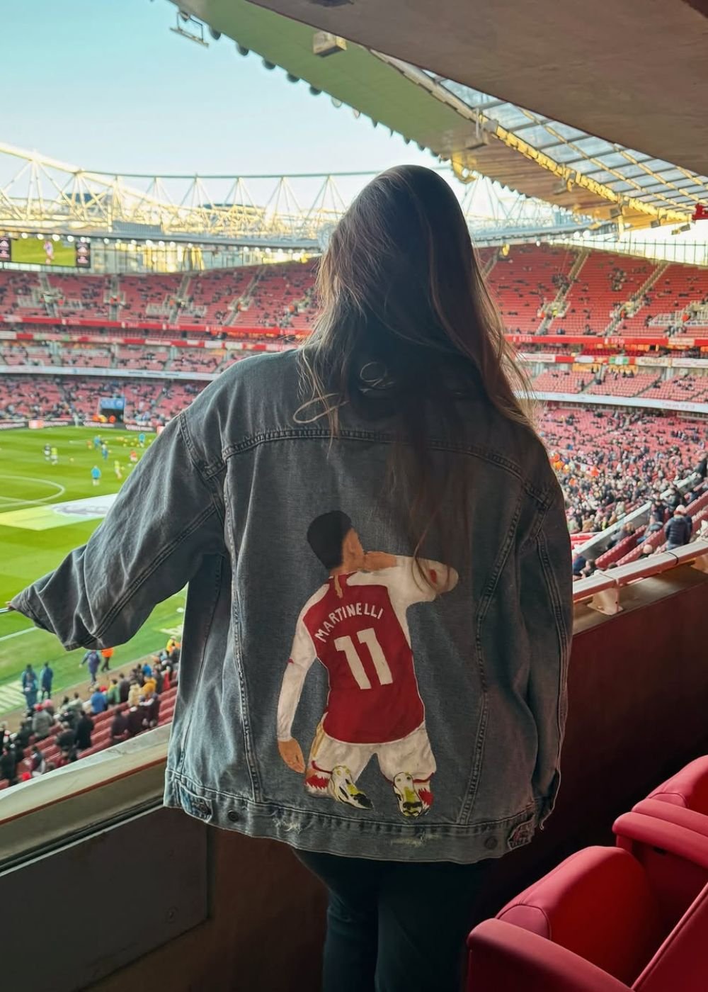 Mulher posa de costas no estádio com jaqueta jeans oversized pintada; vermelho e azul criam clima fã chic.