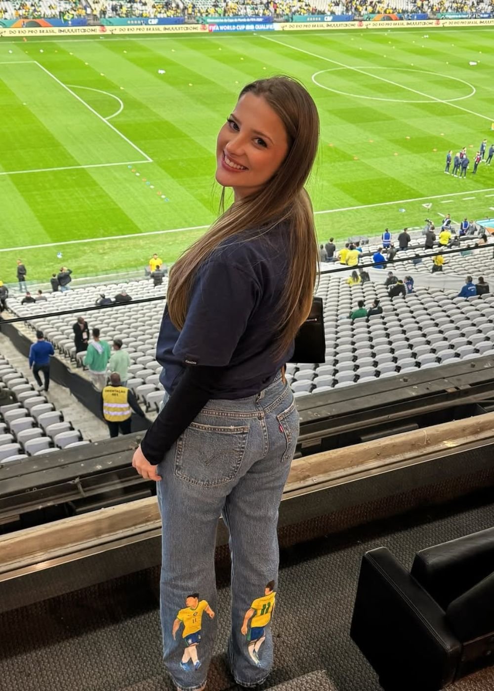 Modelo posa sorrindo no estádio com jeans bordado e camiseta azul-marinho; Afinal, o que é WAG? e porque essas mulheres