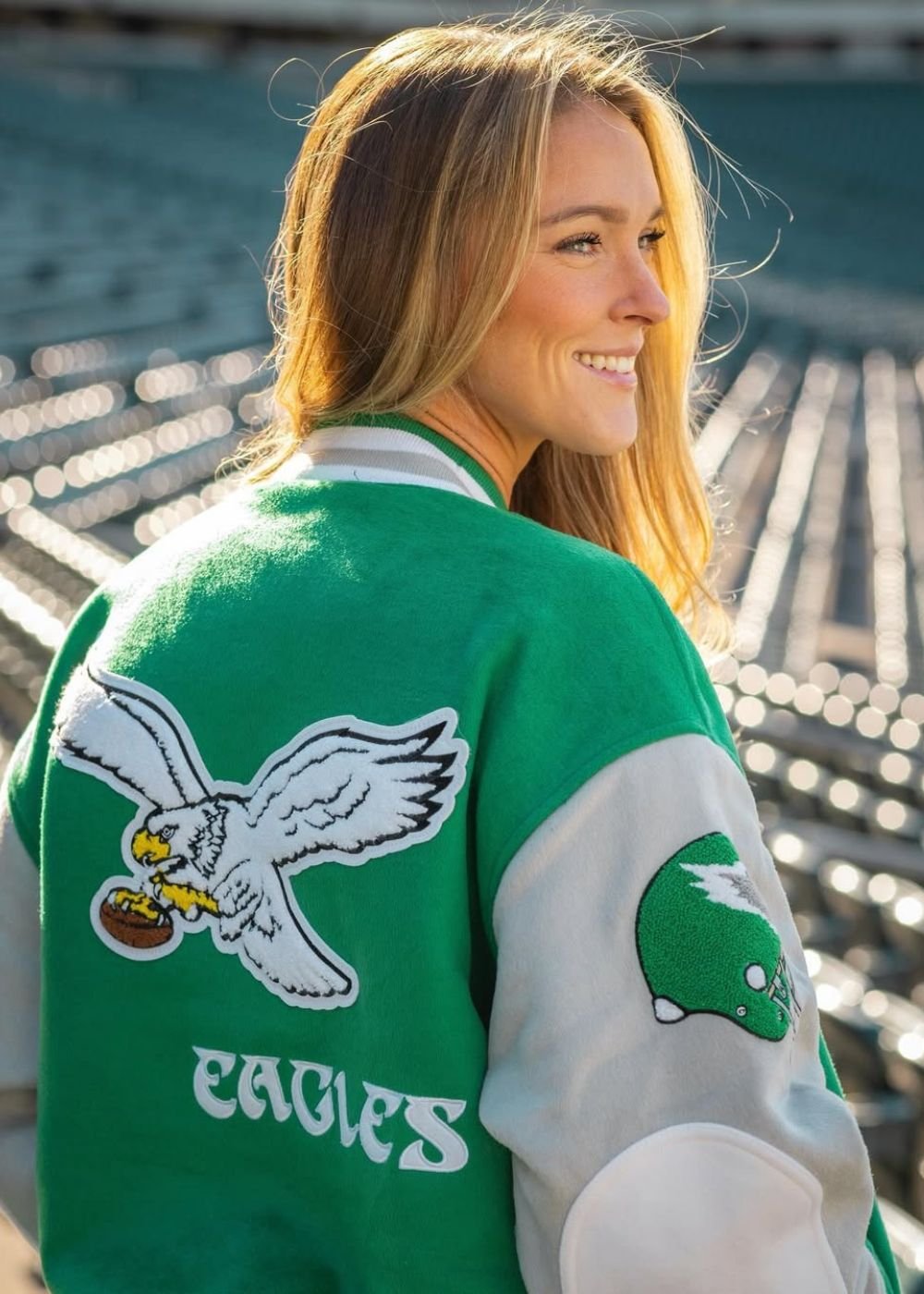 Modelo posa sorrindo em jaqueta varsity verde e branca com patch Eagles em estádio ensolarado; Texto: EAGLES