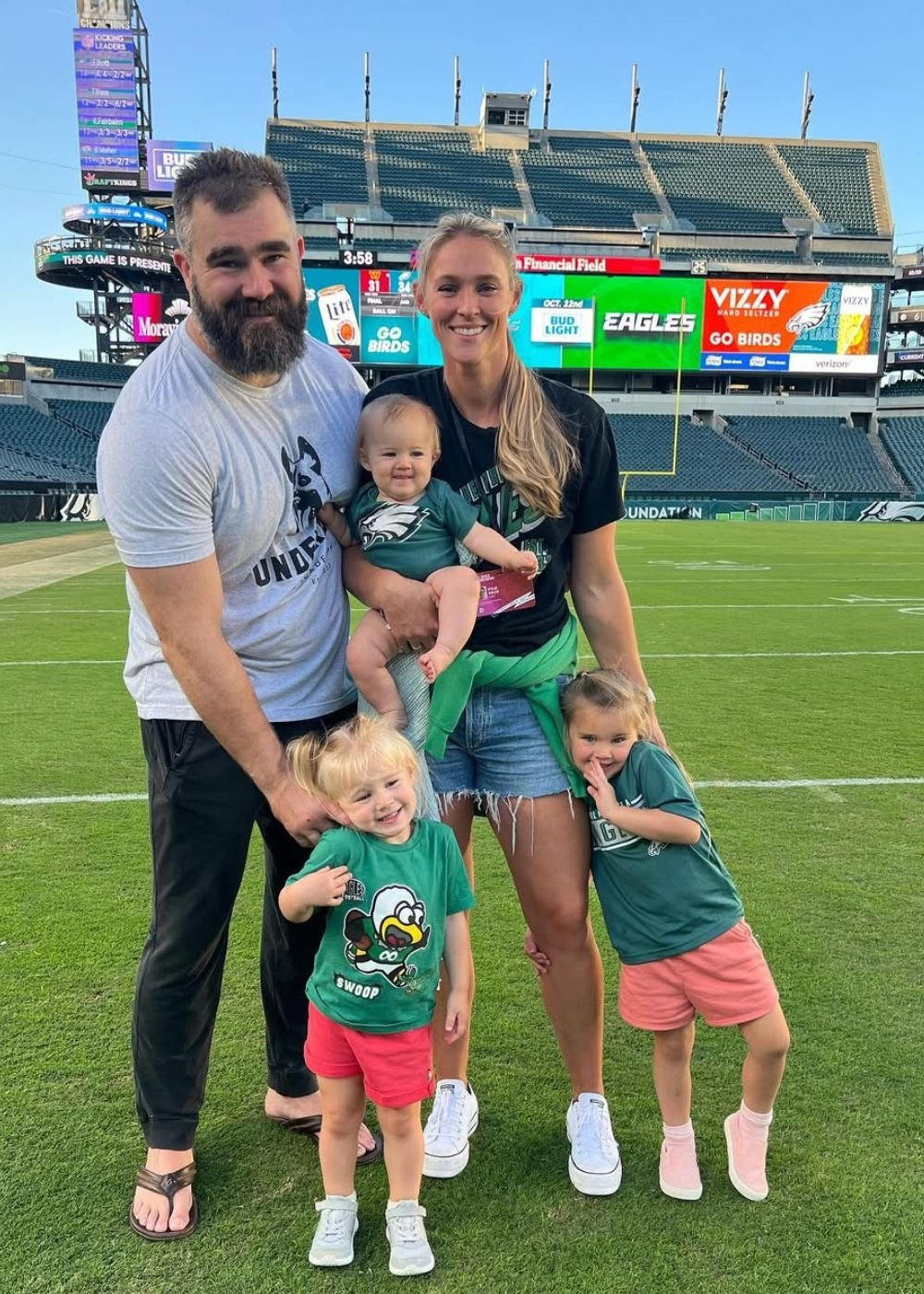 Família posa no gramado com camisetas esportivas verde e preta, jeans e clima afetuoso; Texto: EAGLES GO BIRDS VIZZY BUD
