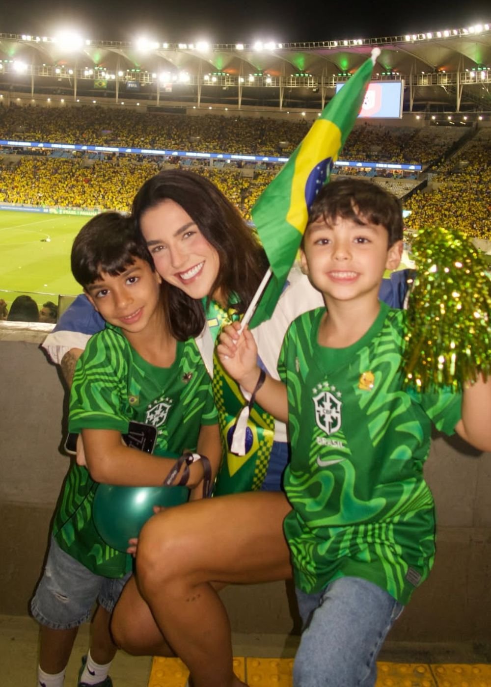 Mulher posa com crianças em camisas verdes do Brasil no estádio lotado, bandeira em destaque e clima vibrante patriótico