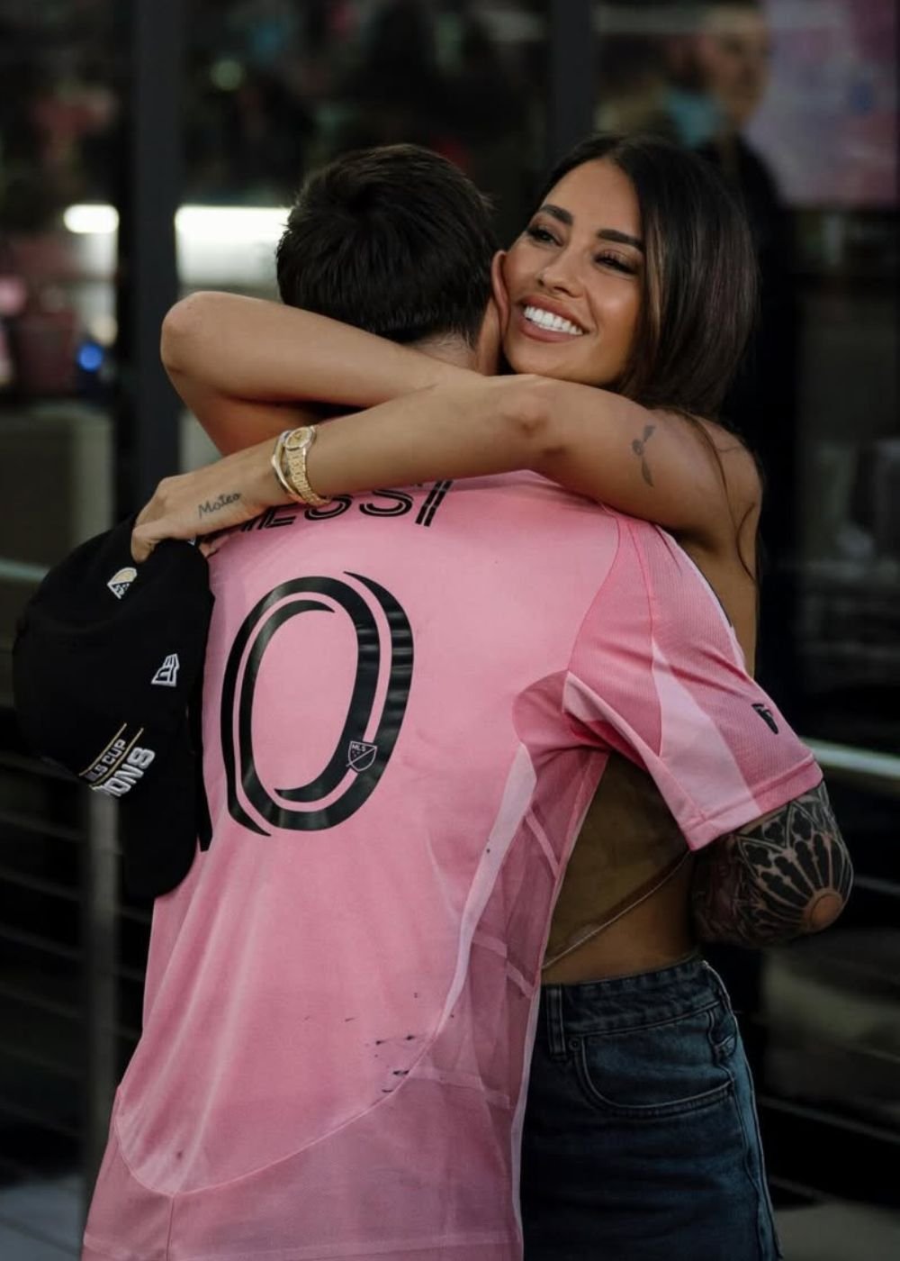 Mulher sorri ao abraçar homem com camisa rosa do Messi; jeans e clima afetuoso. Texto: MESSI 10