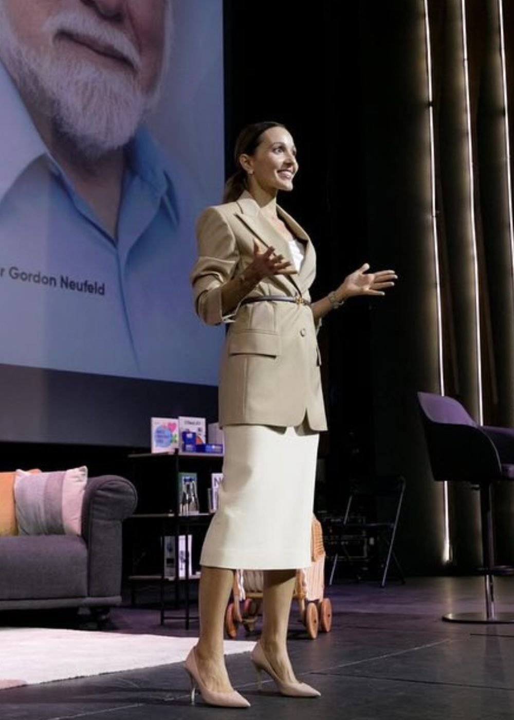 Mulher posa no palco com blazer bege acinturado, saia midi branca e scarpin nude; clima elegante. Texto: Gordon Neufeld