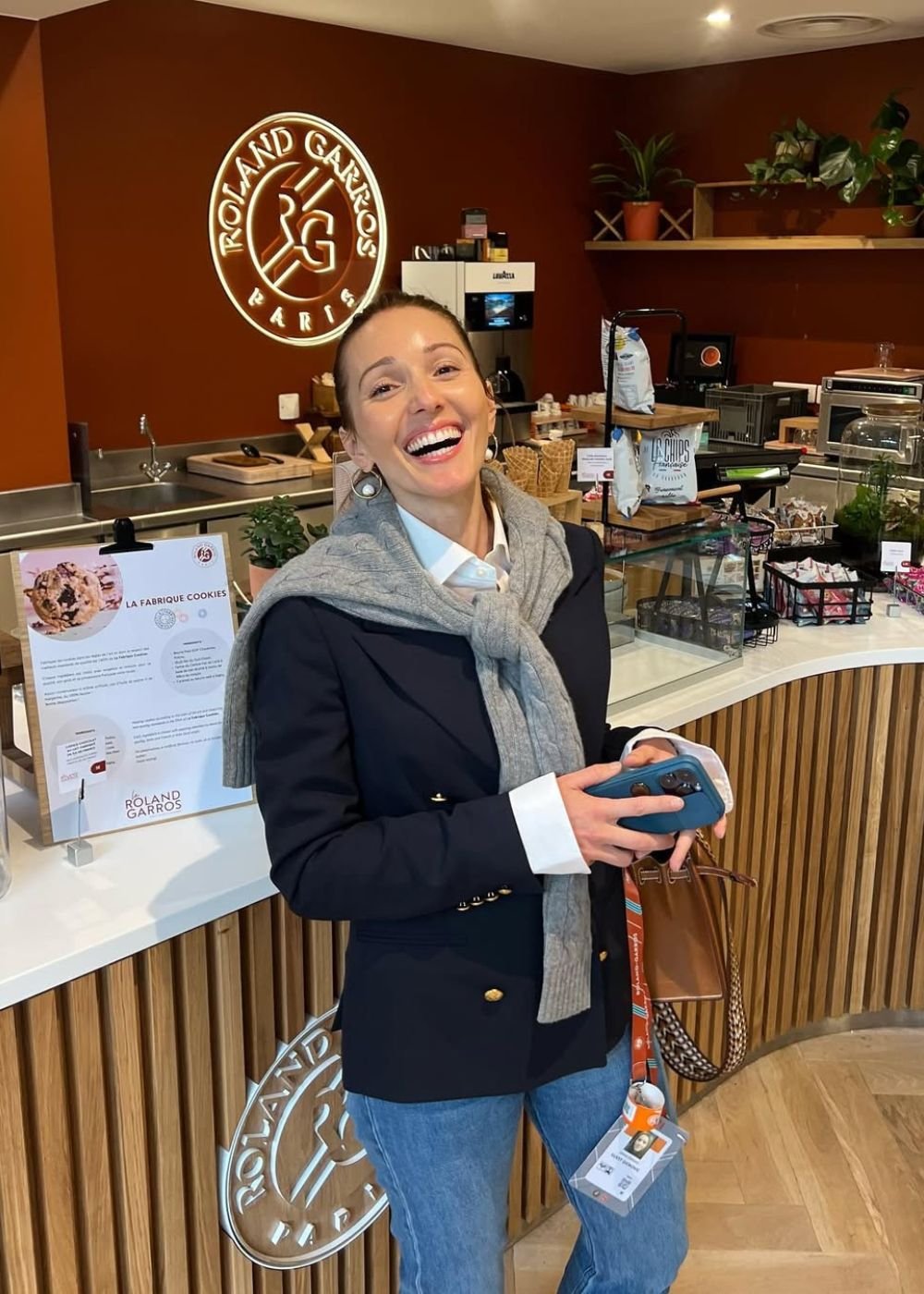 Mulher posa sorrindo com blazer marinho, camisa branca e tricô cinza em café Roland Garros. Texto: ROLAND GARROS PARIS LA