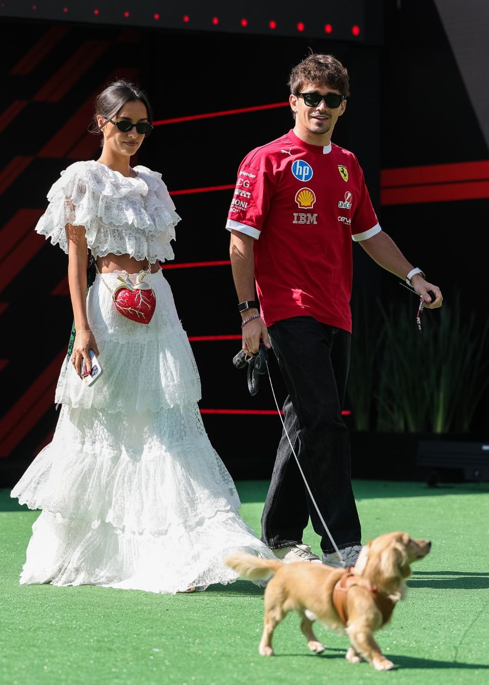 Casal posa com look WAG: vestido branco rendado e camisa Ferrari vermelha, clima glamouroso e descontraído.
