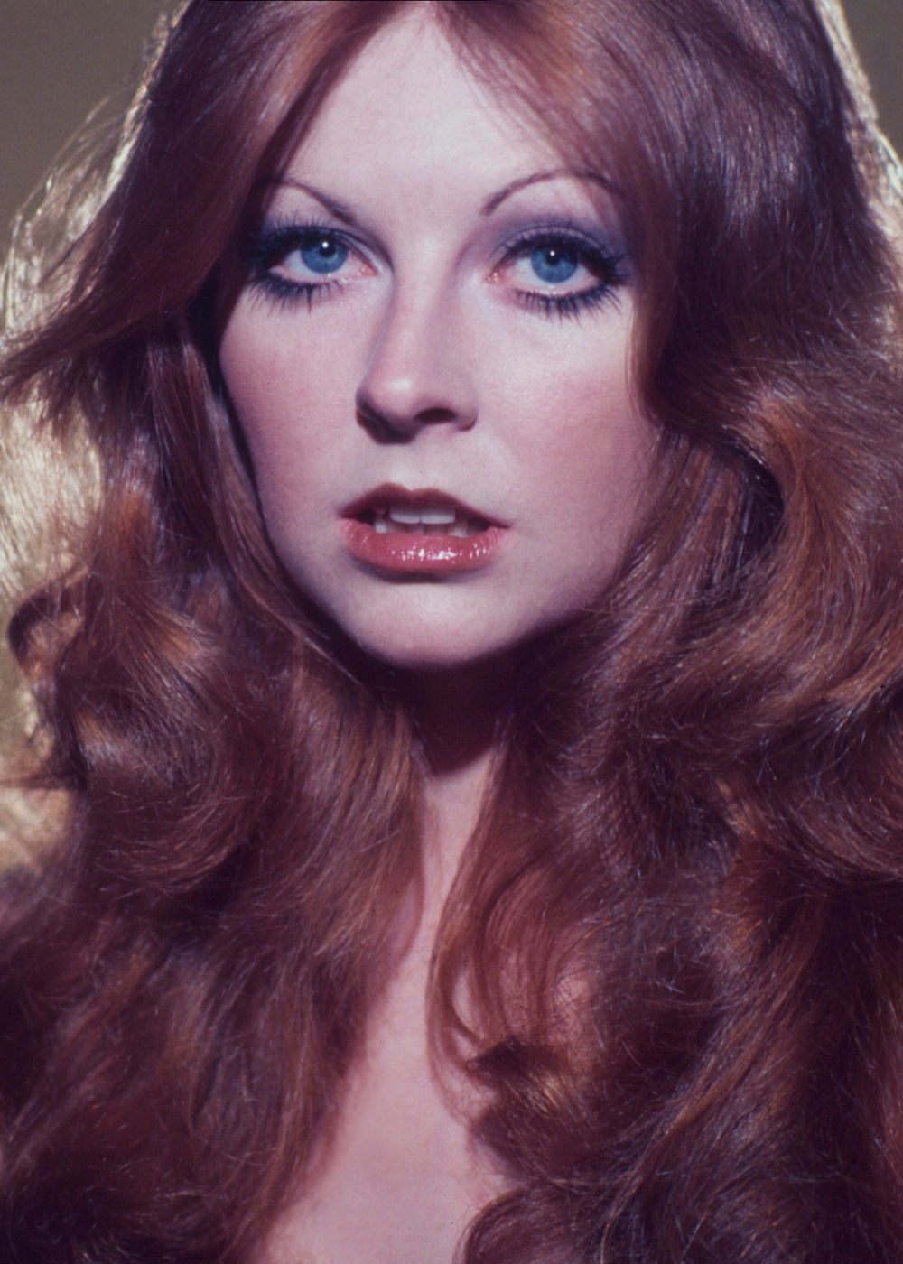 Maquiagem dos anos 70 em close: modelo posa com cílios marcados, sombra suave, lábios brilhantes e cabelo ruivo volumoso.