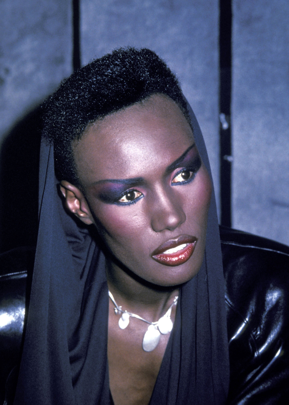 Maquiagem dos anos 80: modelo posa com sombra roxa dramática, couro preto brilhante e colar prateado, clima intenso.