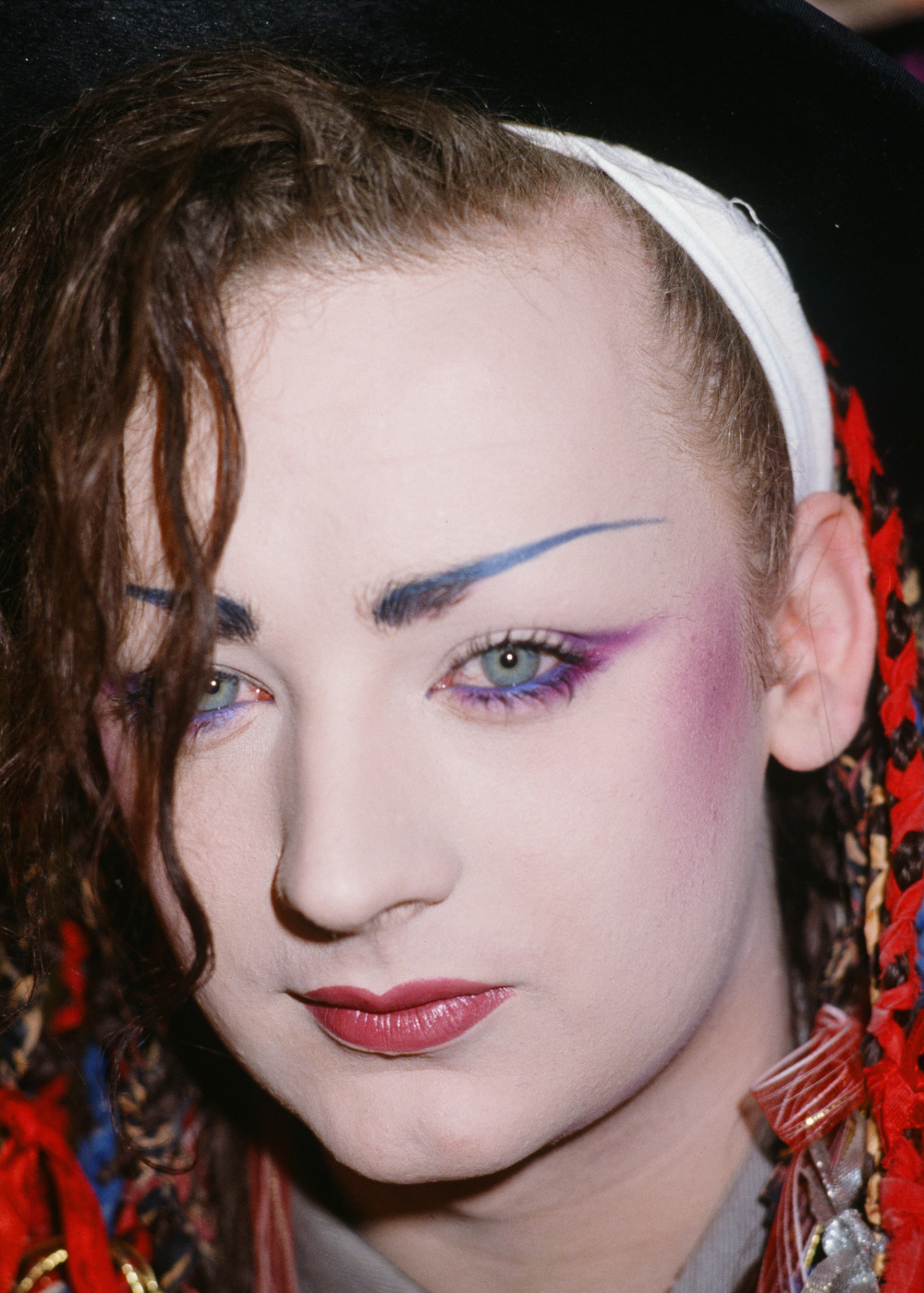Maquiagem dos anos 80 em close: modelo posa com sombra lilás, blush rosado, sobrancelha azul e tranças coloridas.