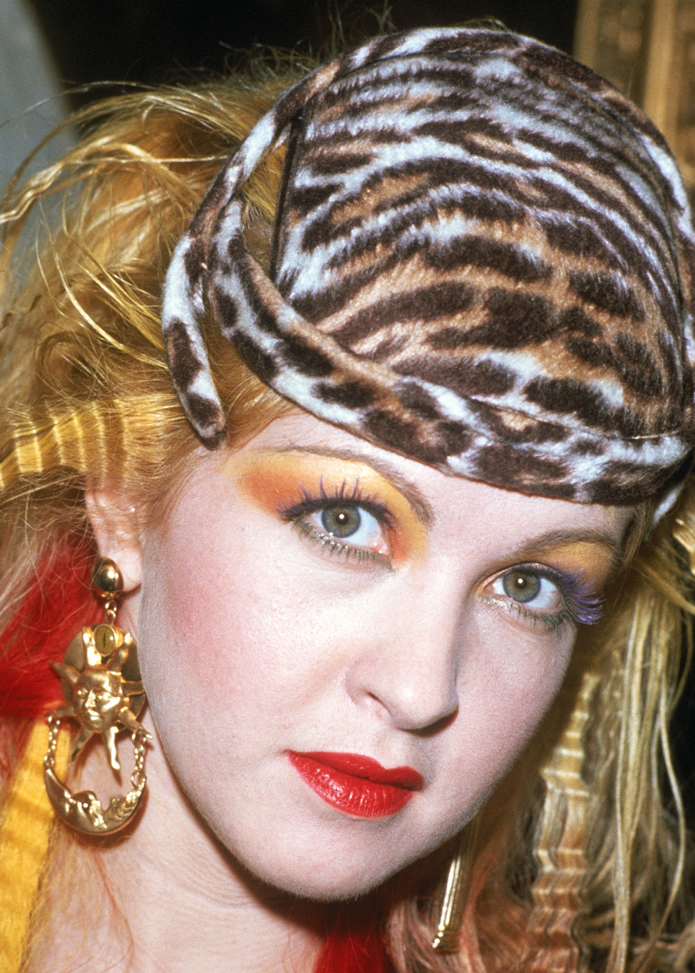 Maquiagem dos anos 80 em close: modelo posa com sombra vibrante, batom vermelho, faixa animal print e maxi brinco